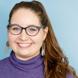 A woman with brown hair, wearing glasses, earrings, and a purple sweater, smiling against a light blue background