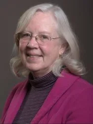 an older woman wearing a pink coat against a grey background