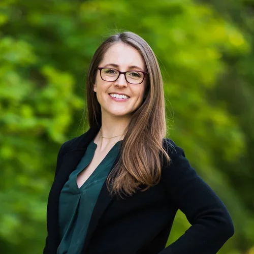 A woman with brown hear and glasses, wearing a black suit jacket and an emerald green shirt, posed against a bright forested background