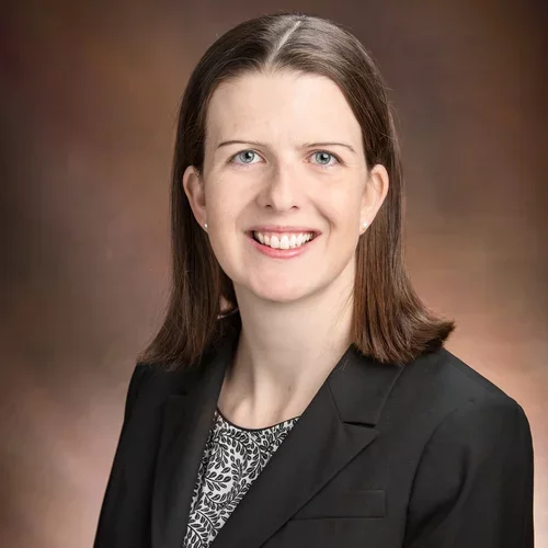 A woman with brown hair and blue eyes, wearing a black suit jacket and speckled shirt, smiling against a professional brown background