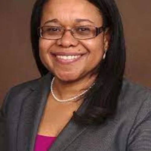 A woman with black hair, wearing a grey suit jacket against a brown background