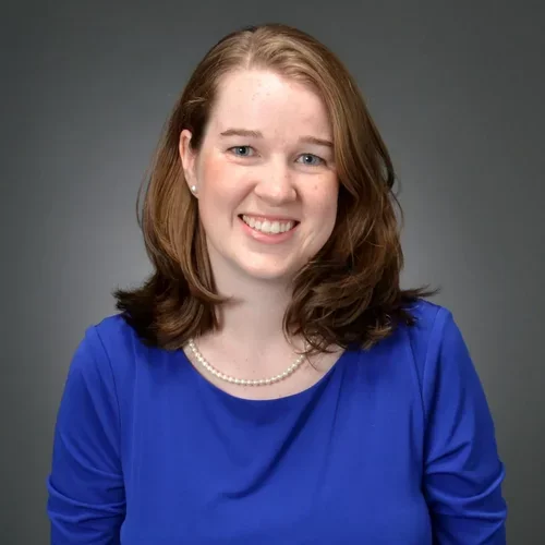 A woman with auburn hair and light eyes, wearing a necklace and royal blue shirt, smiling against a dark background