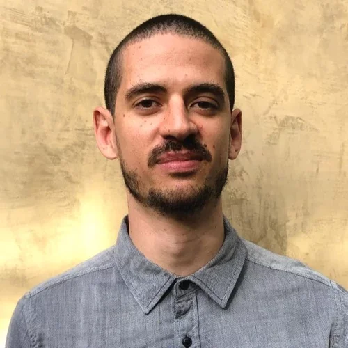 A man with a buzzcut and a beard, wearing a grey collared shirt, smiling against a beige background