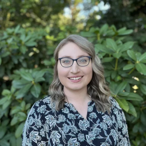A blonde woman smiling, wearing a floral patterned shirt and glasses, against a forested background