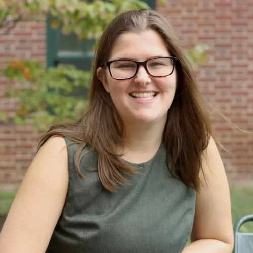 A woman with brown hair and glasses, wearing a dark green dress, seated in front of a brick building in a park