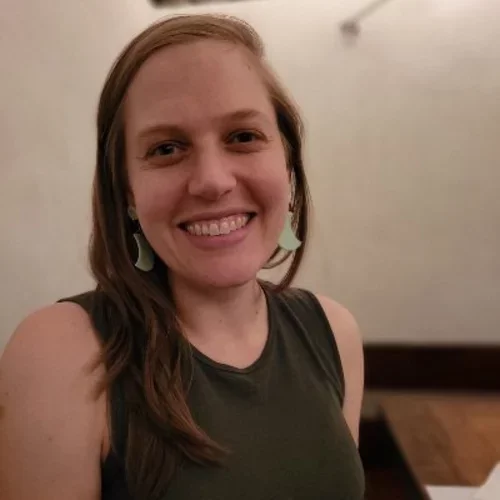 A woman with auburn hair, smiling at the camera, wearing jade earrings and a dark shirt