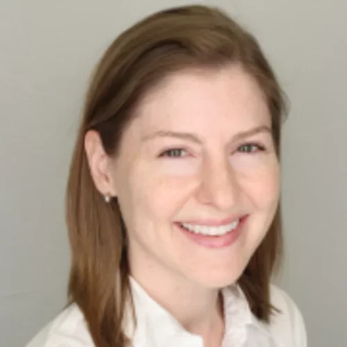 A woman with light brown hair and blue eyes, wearing a white collared shirt, smiling against a beige background