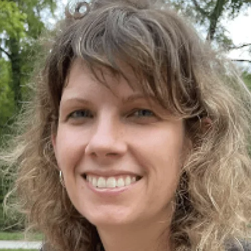 A woman with brown hair wearing a black suit jacket, smiling against a forested background