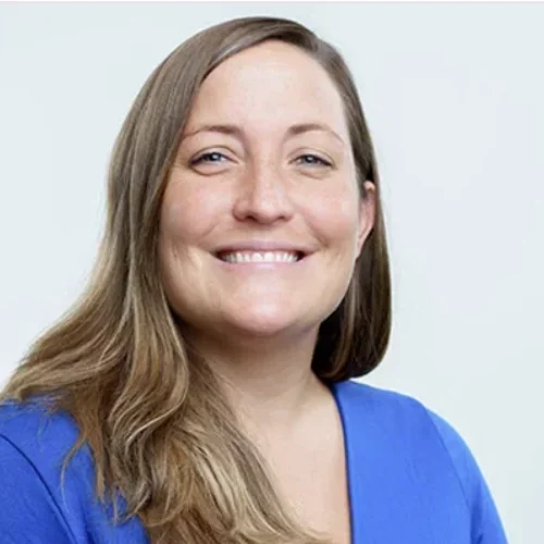 A woman with brown hair, wearing a blue shirt, smiling against a white background