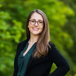A woman with brown hear and glasses, wearing a black suit jacket and an emerald green shirt, posed against a bright forested background