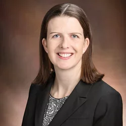 A woman with brown hair and blue eyes, wearing a black suit jacket and speckled shirt, smiling against a professional brown background