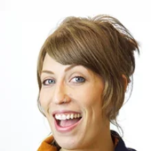 A woman with short brown hair and blue eyes, smiling with her mouth open against a white background