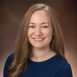 A blonde woman with blue eyes, wearing a blue dress, smiling against a neutral background