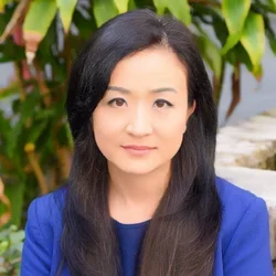 A woman with black hair, wearing a bright blue suit jacket and shirt, seated in front of palms