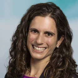 A brunette woman, wearing a black suit jacket with a purple shirt, against a neutral blue photography background