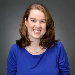 A woman with auburn hair and light eyes, wearing a necklace and royal blue shirt, smiling against a dark background