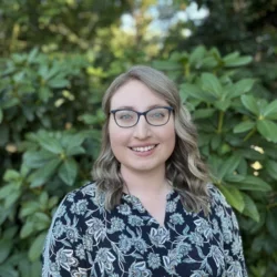 A blonde woman smiling, wearing a floral patterned shirt and glasses, against a forested background