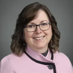 A woman with wavy brown hair, wearing glasses and a light pink overcoat, smiling in front of a grey background