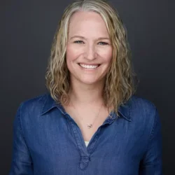 A woman with blonde hair and blue eyes, wearing a blue collared shirt with a necklace, smiling in front of a dark grey background