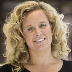 A woman with curly blonde hair and blue eyes, wearing a black shirt, smiling with her head tilted in front of a blurred background