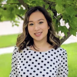 a woman with light brown hair, wearing a white shirt with black polka dots, smiling in front of a forested background