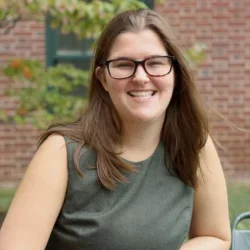 A woman with brown hair and glasses, wearing a dark green dress, seated in front of a brick building in a park