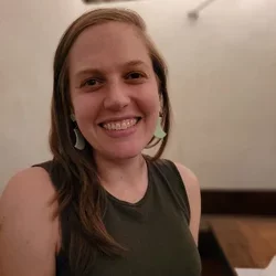 A woman with auburn hair, smiling at the camera, wearing jade earrings and a dark shirt