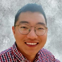 A man with short hair and glasses, wearing a red, white, and blue plaid shirt, smiling against a concrete grey background