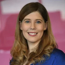A woman with light brown hair and blue eyes, wearing a blue shirt, smiling against a blurred pink background