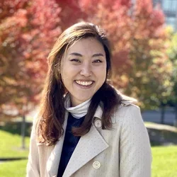 A woman sitting on a park bench, wearing a long brown coat, holding a cup of coffee