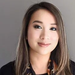 A woman with light brown hair, wearing a scarf with a black shirt, smiling against a dark grey background
