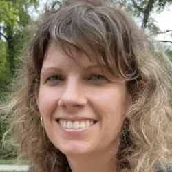 A woman with brown hair wearing a black suit jacket, smiling against a forested background