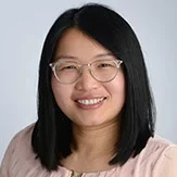 A woman with black hair, glasses, and a pink shirt, smiling against a grey background