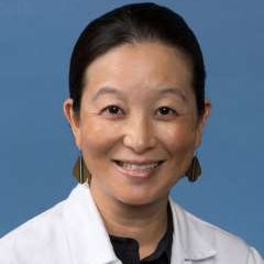 A doctor with black hair and earings, wearing her doctors coat against a blue background.