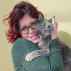 A woman with red hair, glasses, and a green sweater, smiling and posing with her grey speckled cat