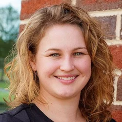 A woman with wavy ginger hair and green eyes, wearing a black shirt, smiling against a brick background